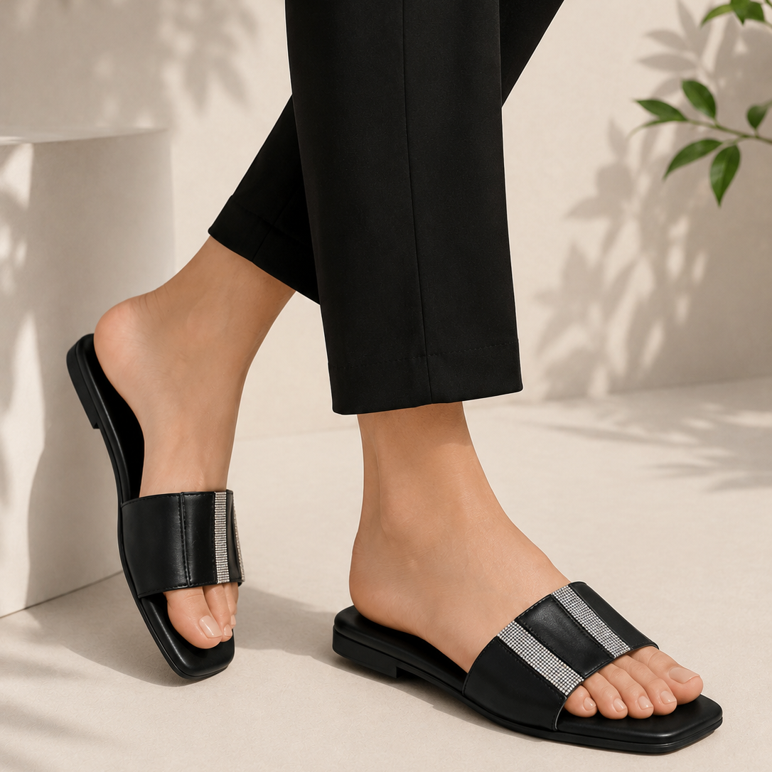 A person wearing black slide sandals with wide metallic silver straps, shown from the calves down in a styled indoor setting with black cropped pants and soft shadow details.